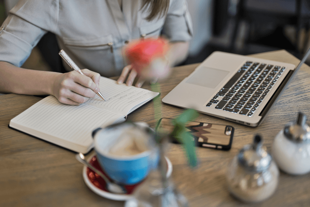 a person writing on a notebook with an open laptop on the side